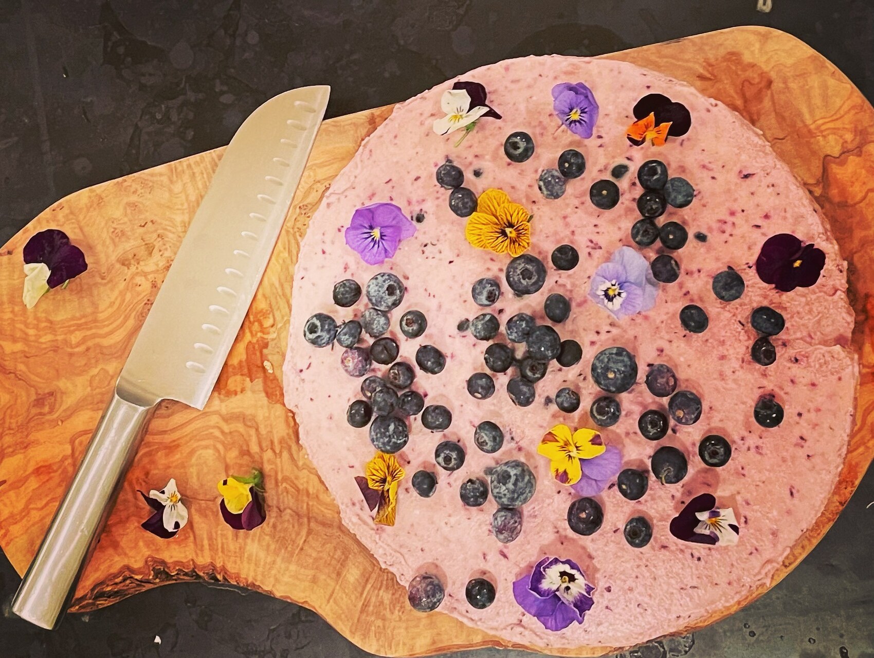 Whole blueberry mousse cake on an olive wood board, scattered with blueberries and purple, yellow and white violas, with a kitchen knife alongside