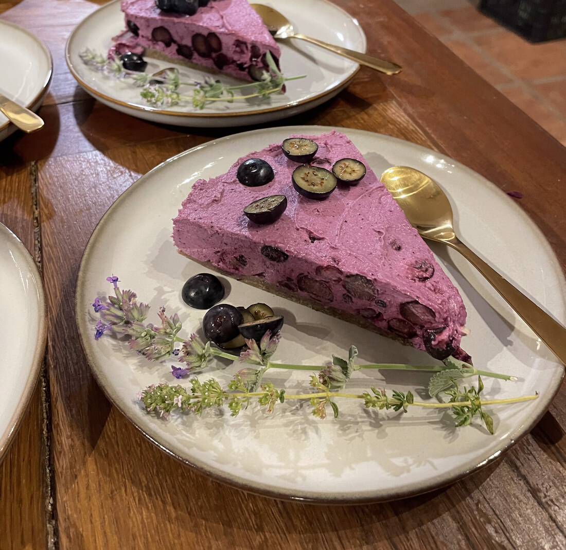 A slice of blueberry mousse cake on a cream plate with a gold spoon, showing whole blueberries through the mousse and a date base, garnished with lavender and thyme sprigs